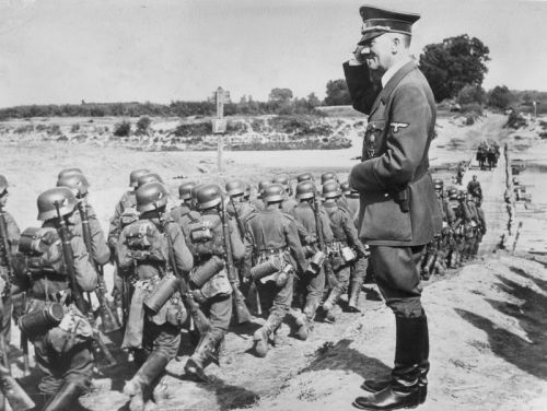 Hitler salutes as he oversees troops during the Nazi occupation of Poland. The troops march in formation toward a wooden bridge, constructed by the Nazis across the San River, near Jarolaw, Poland, Sep 1939